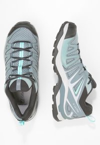 Des chaussures de randonnée grises et teal présentent une tige en mesh, une semelle en caoutchouc et un système de laçage bien visible. Les accents notables incluent une bande turquoise contrastante.