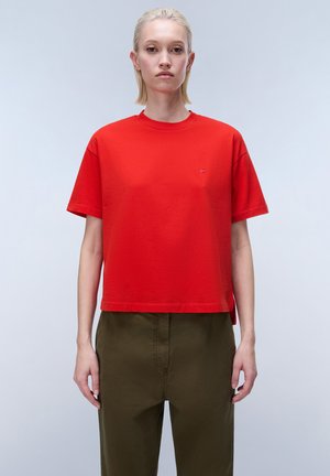 Femme debout, de face, portant un t-shirt rouge vif à manches courtes et un pantalon vert olive sur un fond clair uni.