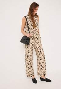 Beige jumpsuit met een bladerenpatroon in bruin en zwart, met een mouwloos ontwerp, losse broekspijpen, gecombineerd met een kleine zwarte handtas en zwarte loafers.