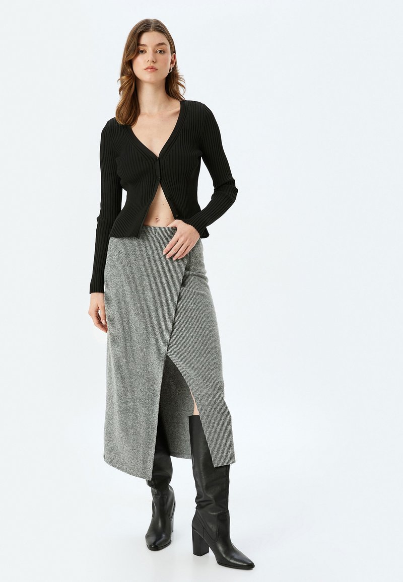 Gilet noir côtelé avec un col en V profond et des boutons, associé à une jupe enveloppante en tricot gris avec une fente à l'avant, portée avec des bottes noires montantes.