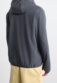 Grå fleecetröja med huva, med en avslappnad passform, elastisk fåll och långa ärmar, kombinerad med gula byxor för kontrast.