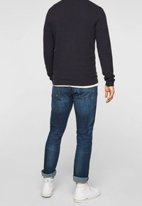 Granatowy sweter o teksturowanej powierzchni z prążkowanym wzorem, w połączeniu z ciemnoniebieskimi, dopasowanymi jeansami oraz białymi, wysokimi trampkami, widoczne podwinięte mankiety.