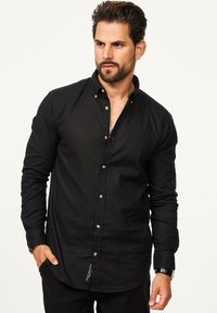 Schwarzes Langarmhemd aus einem Stoff mit glatter Textur, mit einem Button-Down-Kragen, Knopfleiste und einem kleinen Etikett am Saum.