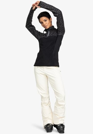 Black fleece jacket with patterned sleeves, zip collar; cream pants, tapered fit; black ski boots. Focus on functional winter sports attire.