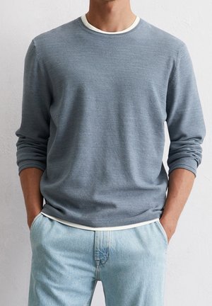 Mann trägt einen strukturierten, hellblauen Langarmpullover über einem weißen Unterhemd mit hellblauen Jeans, Hände in den Taschen vor weißem Hintergrund.