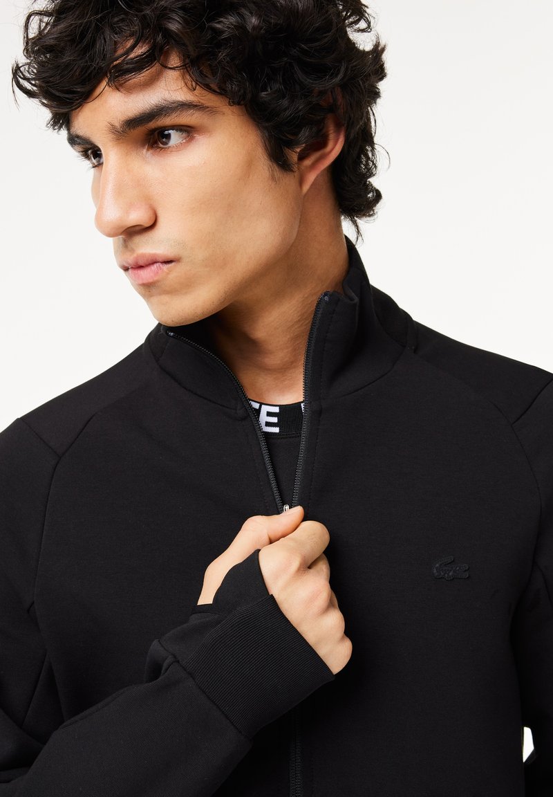 Giacca nera con zip realizzata in tessuto morbido, con colletto alto, maniche aderenti con aperture per i pollici e logo Lacoste sul petto.