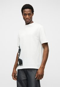 Νεαρός άντρας που φοράει λευκό T-shirt με γραφικά και σκούρο τζιν, στέκεται μπροστά σε απλό ανοιχτό φόντο, κοιτάζοντας προς τα εμπρός με ελαφρύ χαμόγελο.