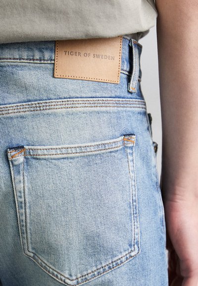 Light blue denim jeans featuring a beige leather patch labeled "TIGER OF SWEDEN" on the back, with subtle fading and orange stitching.