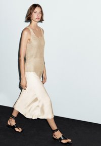 Débardeur beige tricoté, jupe en satin crème fluide et sandales noires à lanières. Textures lisses et coupe décontractée.