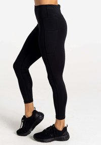 Hoch taillierte schwarze Leggings aus dehnbarem Stoff, mit seitlichen Taschen und glatter Textur für Komfort. Getragen mit schwarzen Sportschuhen.