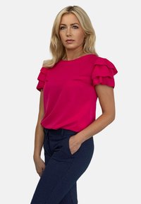 Fuksjowa bluzka z warstwowymi falbanami na rękawach, miękka tkanina, okrągły dekolt, w komplecie z granatowymi spodniami we wzory z bocznymi kieszeniami.