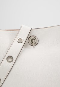 Sac à main en cuir blanc texturé en gros plan comportant une sangle avec des œillets métalliques et un fermoir pivotant circulaire argenté.