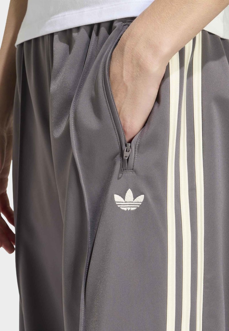 Main dans la poche zippée d'un pantalon de survêtement gris Adidas avec un design à trois bandes blanches et le logo trèfle sur le côté.