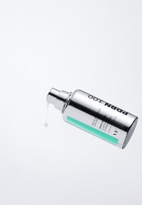 Söljig cylindrisk flaska med pumpdispenser, prydd med ett turkos rand, innehållande en vit vätska, med en liten droppe synlig.