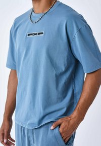 Camiseta azul de manga corta hecha de una tela suave, con un logo rectangular "PXP" en el centro y un diseño de corte holgado.