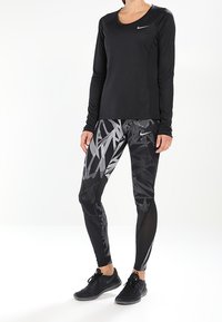 Maglietta sportiva nera con maniche lunghe e scollatura arrotondata; leggings neri con motivi abstract di foglie grigie; scarpe sportive nere.
