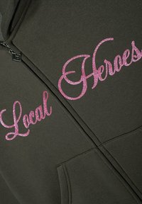 Hoodie wykonany z ciemnozielonego materiału, z różowym brokatowym napisem "Lokalni Bohaterowie" i "Bohaterowie" po lewej stronie. Posiada zamek błyskawiczny oraz przednią kieszeń.