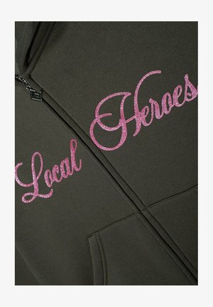 Hoodie wykonany z ciemnozielonego materiału, z różowym brokatowym napisem "Lokalni Bohaterowie" i "Bohaterowie" po lewej stronie. Posiada zamek błyskawiczny oraz przednią kieszeń.