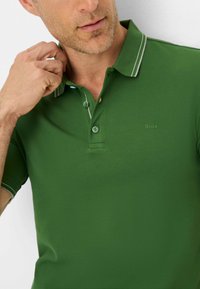 Polo verde realizzato in tessuto testurizzato, dotato di una patta con due bottoni e strisce bianche a contrasto lungo il colletto. Logo del marchio visibile.