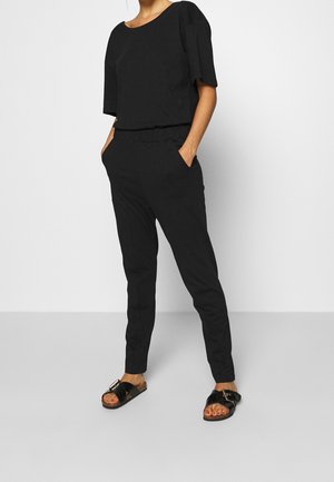 Haut noir à coupe décontractée avec manches courtes et pantalon noir assorti à coupe fuselée. Porté avec des sandales noires ornées d'une boucle décorative.