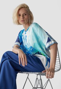 Blaues und weißes Oversized-Oberteil mit abstrakten Mustern, kombiniert mit locker sitzenden blauen Hosen, auf einem Drahtstuhl. Glatte Stoffqualität und entspannte Passform.