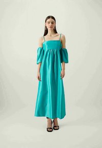 Vestido maxi de cetim aqua com mangas bufantes, decote quadrado e corpete franzido. Combinado com sandálias de salto pretas; modelo em pé contra um fundo neutro.