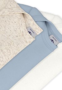 Tři složená trička v béžové, světle modré a bílé barvě. Látky jsou měkké s hladkou texturou; viditelné štítky ukazují velikosti a informace o značce.