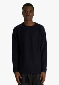 Giovane uomo con indosso un maglione nero a maglia semplice, con maniche lunghe e polsini a costine, in piedi su uno sfondo bianco.