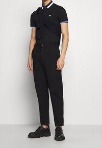 Man med svart pikétröja med blå och vit trim, svarta byxor, svarta finskor och en mörk tröja över axlarna.