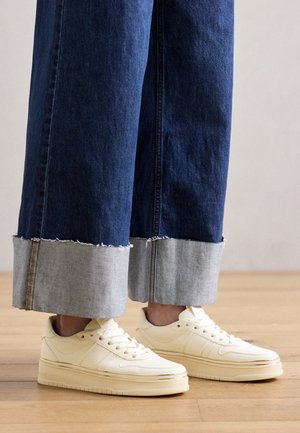Personne portant un jean bleu large avec revers et des baskets basses blanc cassé, debout sur un sol en bois.