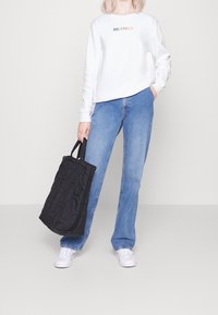 Sudadera blanca con el logo "HILFIGER", jeans azules y un bolso de mano negro acolchado con asas cortas. La modelo lleva zapatillas blancas.