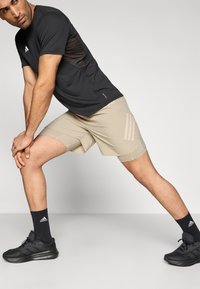 Zwart sportshirt met zijpanelen van mesh, beige korte broek met drie strepen, zwarte sneakers en zwarte sokken. Rekpose tegen een lichte achtergrond.