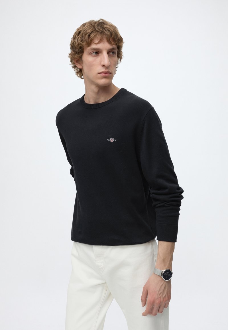 Young man with light curly hair wearing a black long-sleeve shirt and white pants, looking to the side against a plain background.