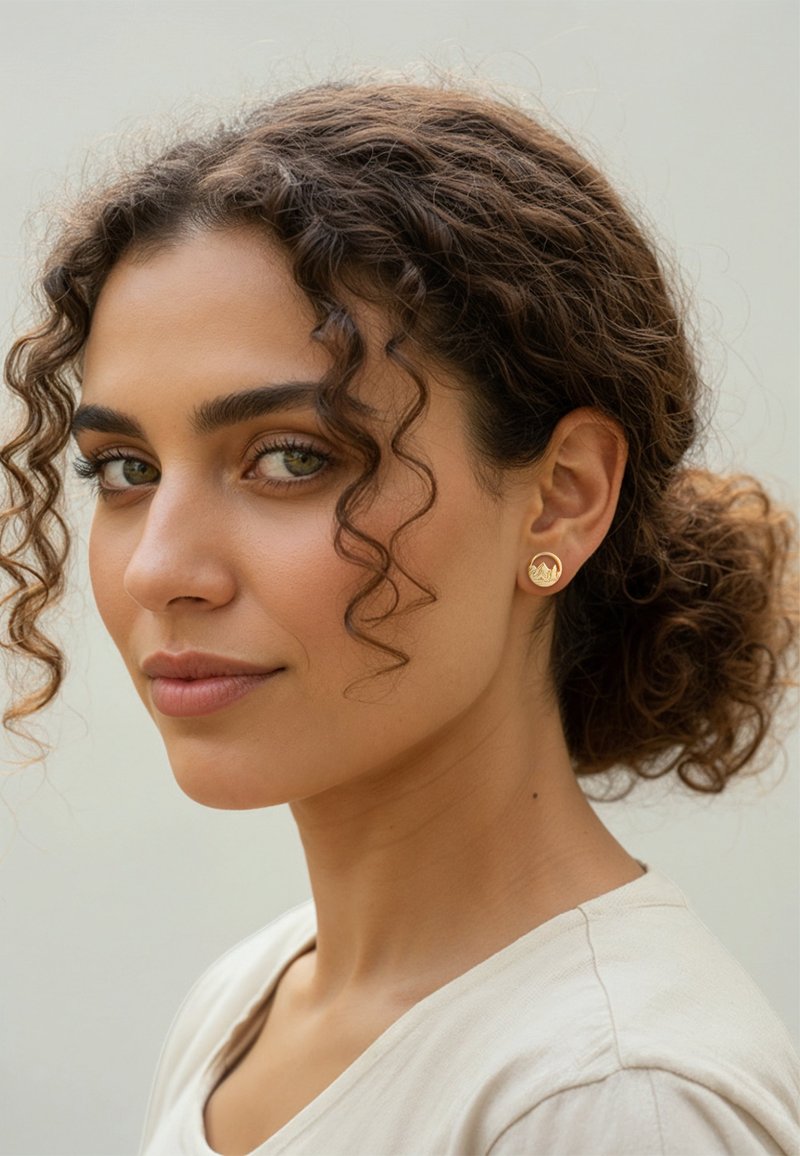 Guldtonade runda örhängen med en bergdesign. Modellen har lockigt hår och bär en ljus top, framför en neutral bakgrund.