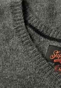 Pull en maille gris avec un col côtelé, présentant une finition texturée et une étiquette noire avec un logo orange.
