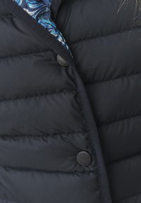 Gros plan sur une veste matelassée noire avec des boutons-pression noirs, portée sur un col de chemise bleu à motifs visible au niveau de l'encolure.