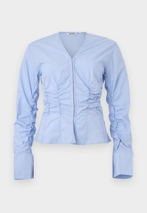 Maglietta a maniche lunghe azzurro chiaro con scollo a V, lati arricciati e vita aderente. Presenta ampi polsini strutturati e un tessuto liscio e leggero.
