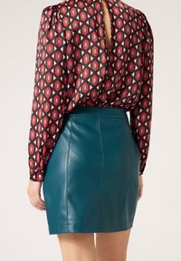Rote und schwarze gemusterte Langarmbluse mit einem V-förmigen Ausschnitt hinten, kombiniert mit einem türkisfarbenen Leder-Minikleid.