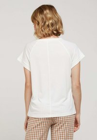T-shirt blanc à manches courtes avec col rond et manches raglan, présentant un détail de couture au centre dans le dos. Assorti à un pantalon à carreaux marron.