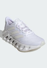 Białe sportowe buty z teksturowaną siateczką u góry, trzema matowymi paskami oraz gumową podeszwą z wyraźnymi krzywiznami i wycięciami.