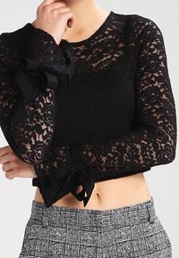 Top in pizzo nero con maniche lunghe, motivo floreale e nastri neri legati ai polsini. Design cropped abbinato a pantaloni a quadri grigi.