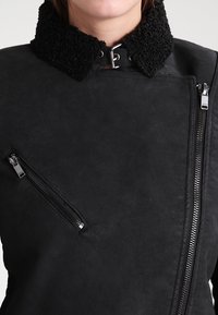 Musta takki pörröisellä mustalla kauluksella, vetoketjulla edessä ja teksturoidulla pinnalla. Sisältää viistosti asetetun vetoketjutaskun ja tyylikkään muotoilun.