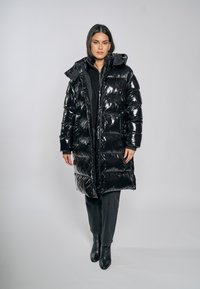 Czarna pikowana kurtka o błyszczącej fakturze, sięgająca do kolan, z kapturem i zamkiem błyskawicznym, noszona na czarnym swetrze i spodniach.