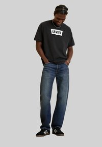 T-shirt en coton noir avec logo Levi's blanc, associé à un jean en denim bleu et des baskets noires avec des rayures blanches. Coupe décontractée et silhouette relaxée.