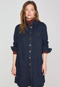 Robe chemise en denim bleu indigo foncé avec des boutons sur le devant, un col, des poches poitrine et des manches retroussées. Présente une coupe décontractée et un ourlet arrondi.