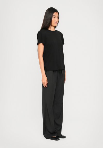 Woman with long black hair wears black short-sleeve top, wide black pants, and black flats, standing sideways against a white background.