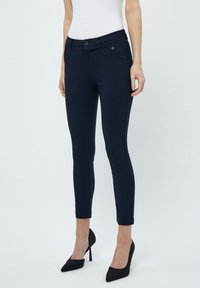 Tmavě modré kalhoty na míru vyrobené z elastického materiálu, s plochým předním dílem, dvěma předními kapsami a slim fit střih. Doplněné černými vysokými podpatky.
