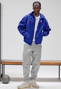 Blå vindjakke med røde detaljer og logo, grå joggebukser og kremfargede joggesko. Modellen står avslappet ved en benk med en basketball.