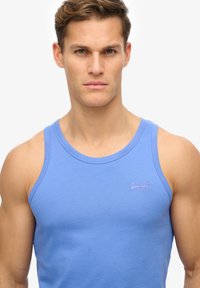Top de algodón azul claro sin mangas con un cuello redondo y tirantes anchos, que presenta un pequeño logo bordado en el pecho. Textura suave.