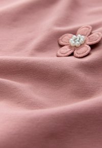 Weiches, leichtes pinkfarbenes Gewebe mit einer strukturierten Blumenapplikation, die in der Mitte mit Perlen verziert ist. Glatte Oberfläche mit dezentem Stretch.